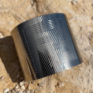 May include: A silver metal cuff bracelet with a geometric pattern. The bracelet is wide and has a polished finish.