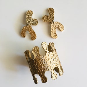 May include: Gold-toned jewelry set with a bracelet and a pair of earrings. The jewelry pieces are made of metal with a textured, organic-shaped design.