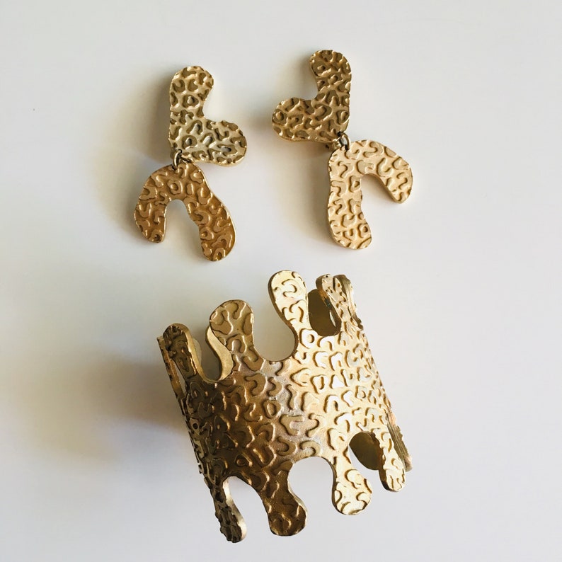 May include: Gold-toned jewelry set with a bracelet and a pair of earrings. The jewelry pieces are made of metal with a textured, organic-shaped design.