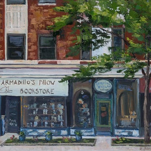 May include: A storefront with a blue awning and the sign "Armadillo's Pillow Bookstore" above the entrance. The storefront has large windows displaying books and other merchandise. A tree with green leaves is in front of the store.