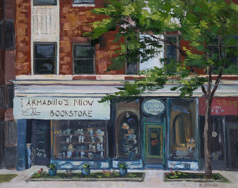 May include: A storefront with a blue awning and the sign "Armadillo's Pillow Bookstore" above the entrance. The storefront has large windows displaying books and other merchandise. A tree with green leaves is in front of the store.