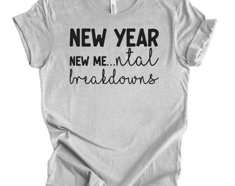 Op de afbeelding: Lichtgrijs t-shirt met de zwarte tekst "NEW YEAR, NEW ME... mental breakdowns". De tekst is in vette en cursieve letters. Het t-shirt heeft korte mouwen en een ronde hals.