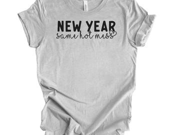 Op de afbeelding: Een lichtgrijs t-shirt met de tekst "NEW YEAR same hot miss" in zwart schrift. Het shirt is gemaakt van een zacht materiaal en heeft korte mouwen. De tekst staat in het midden van de voorkant van het shirt.
