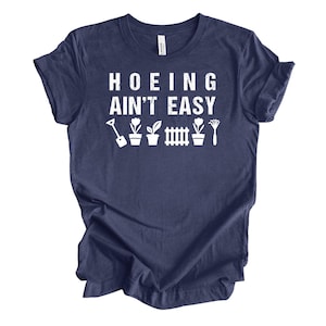 Op de afbeelding: Marineblauwe t-shirt met de witte tekst "Hoeing Ain't Easy". Daaronder witte illustraties van tuingereedschap en planten. Het shirt heeft een ronde hals en korte mouwen.