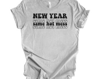 Op de afbeelding: Lichtgrijs t-shirt met de tekst "NEW YEAR" boven "same hot mess" driemaal herhaald in zwart. Het t-shirt heeft korte mouwen en is gemaakt van een zacht materiaal.