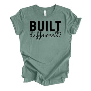 Könnte beinhalten: Ein salbeigrünes T-Shirt mit dem schwarzen Aufdruck "BUILT different". Das Wort "BUILT" ist fett und ohne Serifen, während "different" in Schreibschrift gehalten ist. Das T-Shirt hat kurze Ärmel und einen Rundhalsausschnitt.