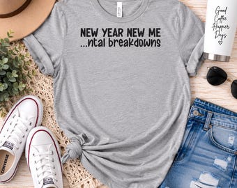 Op de afbeelding: Een grijs gemêleerd T-shirt met korte mouwen en de zwarte tekst "NEW YEAR NEW ME ...ntal breakdowns" op de voorkant. Het shirt wordt getoond naast een beige hoed, een witte beker met zwarte tekst, een zonnebril en een spijkerbroekshort.
