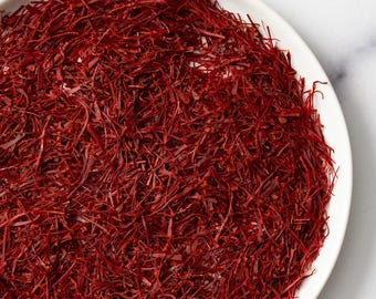 May include: A white ceramic plate filled with vibrant red saffron threads. The threads are densely packed, creating a rich, textured appearance. The saffron is a deep, reddish-brown color, contrasting with the white plate.