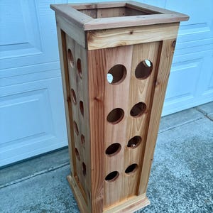 Op de afbeelding: Een hoge, rechthoekige houten plantenbak met een natuurlijke afwerking. De plantenbak heeft meerdere cirkelvormige uitsparingen aan de zijkanten en een vierkante opening aan de bovenkant. De houtnerf is zichtbaar.