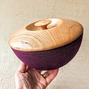 May include: A wooden bowl with a lid and a burgundy fabric lining. The bowl is made of light wood with a natural finish. The lid has a small knob on top.