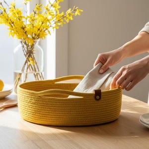 May include: A round, mustard-yellow rope basket with handles, holding a folded linen cloth. The basket has a brown leather accent. A vase of yellow flowers and lemons on a plate are in the background.