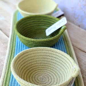 Op de afbeelding: Drie geweven touwkommen in verschillende maten en kleuren. De kommen zijn groen, lichtgroen en beige. Ze zijn gestapeld op een gestreept kleed.