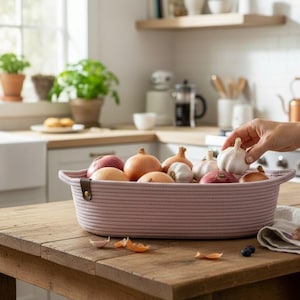 May include: A woven pink basket filled with various fresh produce, including onions, garlic, and a red apple, sits on a wooden table. A hand reaches for a garlic bulb. The kitchen setting includes a window and potted plants.