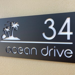 Tropical style house entry sign in matte black with white letters and numbers. Sign has 2 palm trees on the top left, the house number is on the right side with the street name below, a horizontal line separates the numbers and street name.