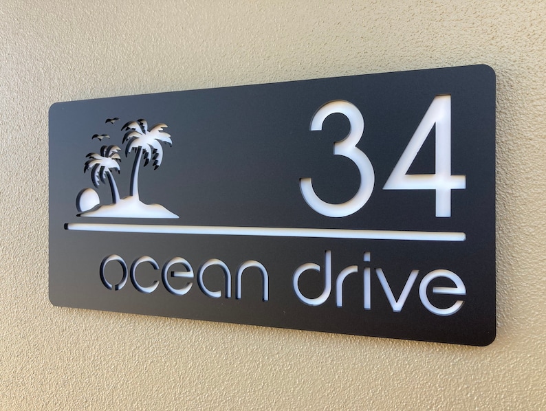 Tropical style house entry sign in matte black with white letters and numbers. Sign has 2 palm trees on the top left, the house number is on the right side with the street name below, a horizontal line separates the numbers and street name.