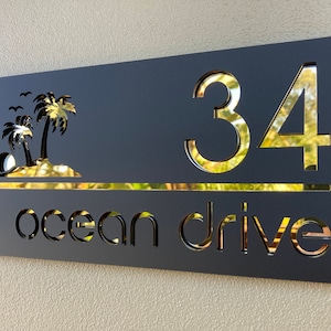 Tropical style house entry sign in matte black with gold mirror letters and numbers. Sign has 2 palm trees on the top left, the house number is on the right side with the street name below, a horizontal line separates the numbers and street name.