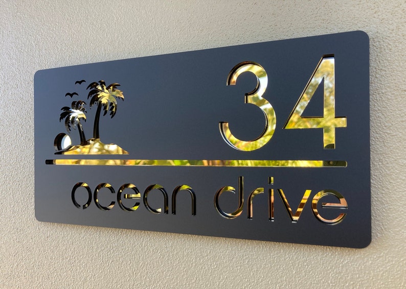 Tropical style house entry sign in matte black with gold mirror letters and numbers. Sign has 2 palm trees on the top left, the house number is on the right side with the street name below, a horizontal line separates the numbers and street name.