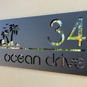 Tropical style house entry sign in matte black with black mirror letters and numbers. Sign has 2 palm trees on the top left, the house number is on the right side with the street name below, a horizontal line separates the numbers and street name.