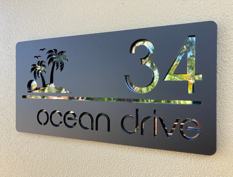 Tropical style house entry sign in matte black with black mirror letters and numbers. Sign has 2 palm trees on the top left, the house number is on the right side with the street name below, a horizontal line separates the numbers and street name.