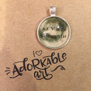 May include: A silver pendant with a glass circle featuring a black and white image of the Sorting Hat from the Harry Potter series. The text "Sorting Hat" is visible on the image. The pendant is on a brown background with the text "i love adorable art" written in black.
