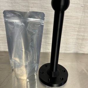 May include: A black 3D printed stand with a round top and a base with six holes. The stand is packaged in a clear plastic bag.