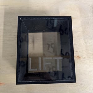 May include: A rectangular black frame with a transparent center panel. The word LIFT is etched in capital letters across the bottom portion of the clear panel. The frame has a clean, sharp-edged design with a smooth finish and a hollow center.