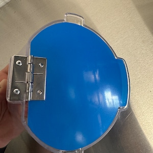 Op de afbeelding: Een doorzichtige plastic container met een blauw deksel. Het deksel heeft een scharnier en is bevestigd aan de container.