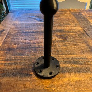 May include: A black metal hook with a round top and a base with multiple holes. The hook is mounted on a wooden surface.