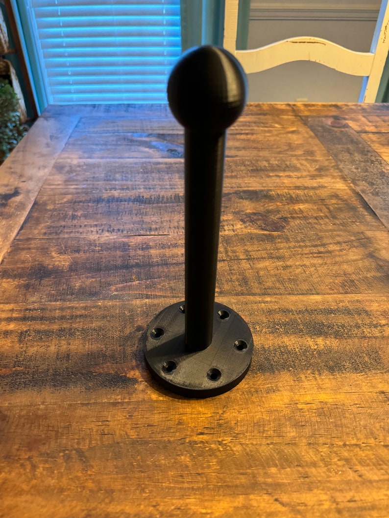 May include: A black metal hook with a round top and a base with multiple holes. The hook is mounted on a wooden surface.