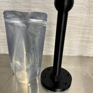 May include: A black plastic 3D printed stand with a round top and a base with six holes. The stand is packaged in a clear plastic bag.