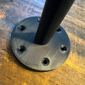 May include: Black metal base with six screw holes for mounting a table leg. The base is round and has a textured surface.