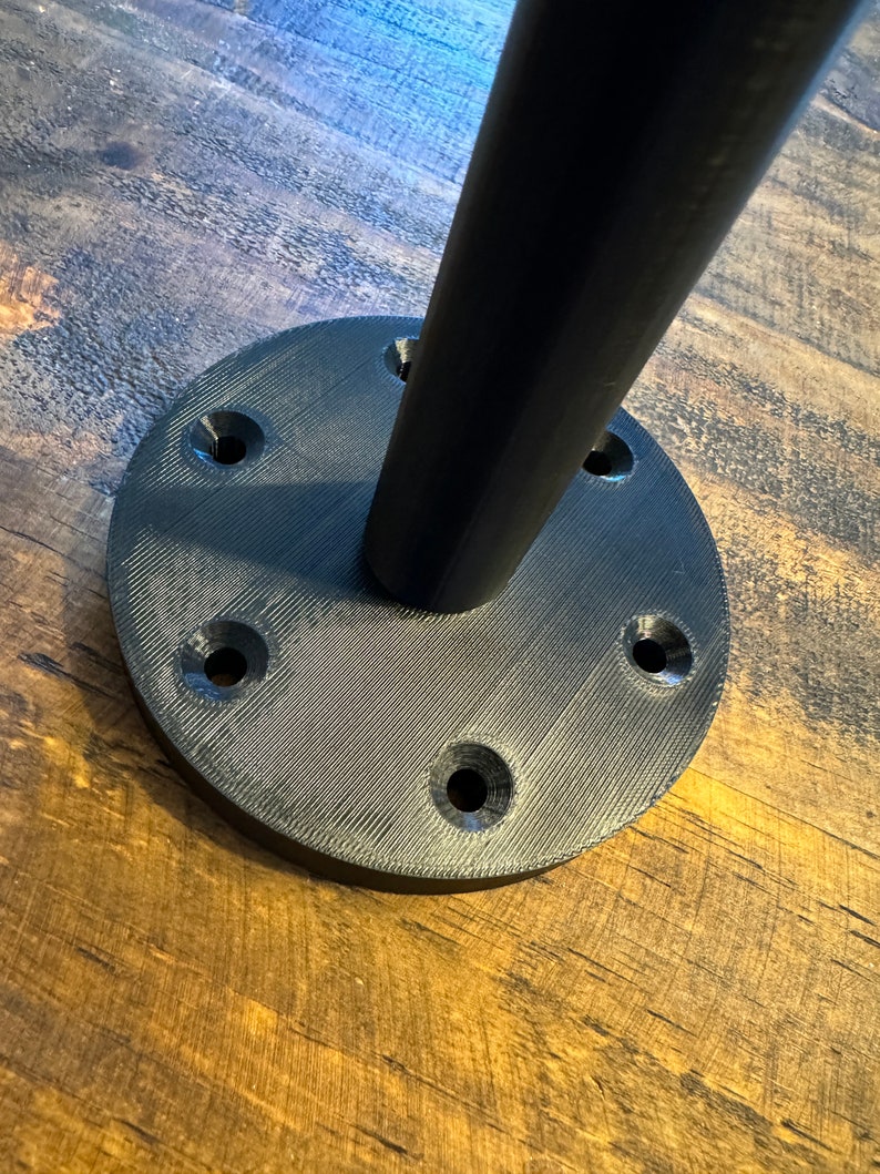 May include: Black metal base with six screw holes for mounting a table leg. The base is round and has a textured surface.