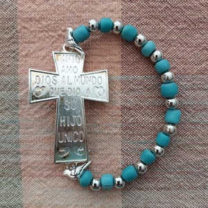 May include: A silver cross charm with the inscription "Tanto amo Dios al mundo que dio a su hijo unico" hangs from a teal blue beaded bracelet.