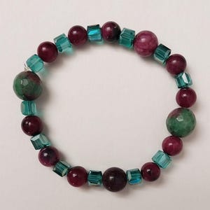 May include: A beaded bracelet featuring alternating burgundy and teal-colored beads. The bracelet includes faceted teal square beads, burgundy round beads, and two larger green and burgundy round beads. The bracelet is displayed on a white surface.
