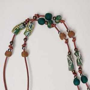 May include: A double-strand necklace with green, brown, and white beads. The necklace is made of leather cord and features a variety of shapes and sizes of beads.