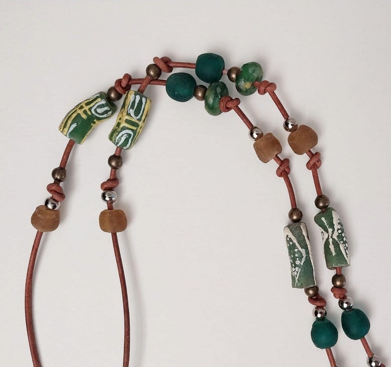 May include: A double-strand necklace with green, brown, and white beads. The necklace is made of leather cord and features a variety of shapes and sizes of beads.