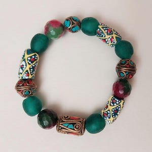 May include: A beaded bracelet featuring a mix of green, red, and gold beads. The bracelet includes faceted red and green beads, dark green round beads, and decorative gold-toned beads with turquoise and red accents. The bracelet is on a light background.