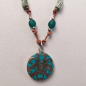 May include: A turquoise and silver pendant necklace with a leather cord and green glass beads. The pendant is round and has a detailed design.