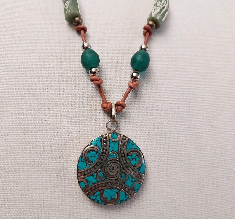 May include: A turquoise and silver pendant necklace with a leather cord and green glass beads. The pendant is round and has a detailed design.