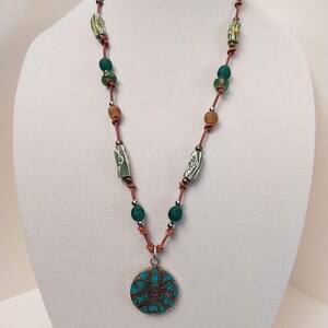 May include: A long necklace with a turquoise pendant. The necklace is made of brown leather cord and features green and brown beads. The pendant is a round, silver-toned disc with a turquoise inlay and intricate designs.