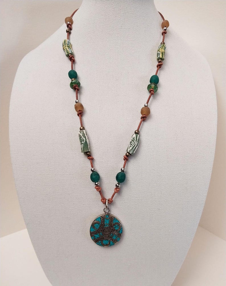 May include: A long necklace with a turquoise pendant. The necklace is made of brown leather cord and features green and brown beads. The pendant is a round, silver-toned disc with a turquoise inlay and intricate designs.