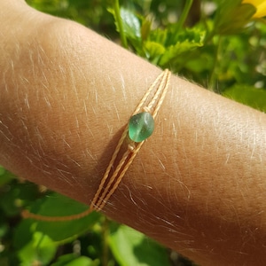 May include: A bracelet featuring a single, smooth, translucent green sea glass bead centred on a triple-strand light tan cord. The cord is braided and knotted to secure the bead in place, creating a simple, minimalist design that wraps around the wrist.