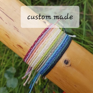 May include: A close-up of a collection of custom-made braided bracelets in various colors, including red, orange, yellow, green, blue, and black. The bracelets are arranged in a rainbow pattern and are displayed on a wooden surface. The text "custom made" is written on a white label above the bracelets.