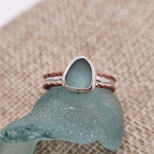 May include: A ring featuring a smooth, frosted light blue sea glass center set in a silver bezel. The band is composed of three stacked layers, with a central silver wire flanked by two textured, copper-colored twisted metal wires.