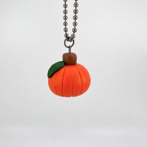 May include: A small orange pumpkin with a green leaf and brown stem hangs from a silver chain. The pumpkin is a handmade charm necklace.