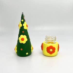 May include: A green cone-shaped bottle stopper with yellow and orange flowers and orange dots. The stopper is attached to a silver chain. A clear glass bottle with a yellow and orange flower design is also shown.