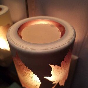 May include: White ceramic electric wax warmer with a leaf design. The warmer has a round opening at the top with a melted wax disc inside. The warmer is lit and glowing.
