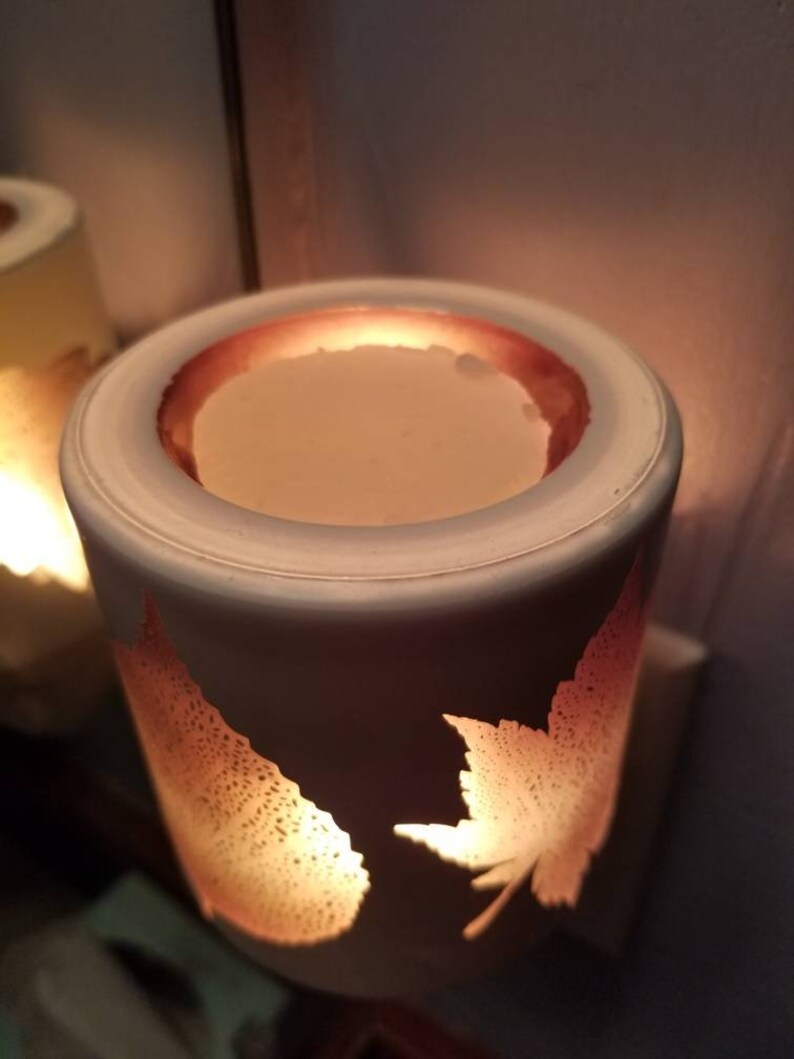 May include: White ceramic electric wax warmer with a leaf design. The warmer has a round opening at the top with a melted wax disc inside. The warmer is lit and glowing.