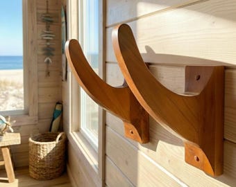 May include: Two curved, light brown wooden wall-mounted brackets with a smooth finish. Each bracket features a wide, flat base plate secured to the wall with visible screws, extending outward into a sweeping, upward-curving support arm.