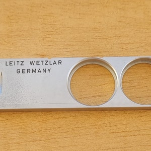 Op de afbeelding: Een zilverkleurig Leitz Wetzlar Germany cameraonderdeel met twee cirkelvormige openingen. De tekst "LEITZ WETZLAR GERMANY" staat bovenaan gedrukt. Het onderdeel heeft een rechthoekige vorm met een klein blauw-wit ontwerp aan de linkerkant.